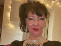 MaitresseElsa with messy brown hair, clear glasses, red lipstick, multiple dangling earrings, and a chunky gold necklace, wearing a black top, looking slightly annoyed.