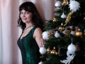 AngieGreen with dark brown hair, green dress, and necklace stands beside a Christmas tree with white and gold ornaments. Her tits are visible under the dress.