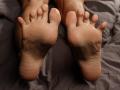 LettieSweet's soles are in focus, showing her bare feet with slightly curled toes and smooth skin, resting on a gray bedsheet. Her toes are slightly spread apart.