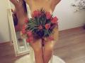 Azazelle's bare ass, covered by a bouquet of red tulips wrapped in brown paper, sitting on a white chair in front of a mirror.