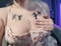 Rubbi's tits have black tattoos of dark, branching shapes above them. She's wearing a floral dress with a spaghetti strap, and her hand covers part of her tits.
