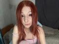 HannaCool with red hair, freckles, and piercings, wearing a pink top, sitting on a bed, looking at the camera, her pink-striped knee visible in the foreground.