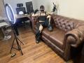 AmySpider with torn black clothes, mask, gloves, and boots, sitting on brown leather couch, holding a gun, ring light on tripod, wooden floor, AmySpider's tattooed chest and legs visible.