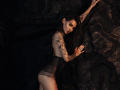 SophieKitty with tattoos, wearing sheer black lingerie, leans against dark rocks, arms raised, looking at camera, showing her ass, with long dark hair and a serious expression.