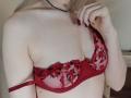 SweetLadyXX wearing a red, sheer bra with black floral embroidery, her hand with purple nail polish touching her collarbone, her pale skin and small to medium-sized tits visible.
