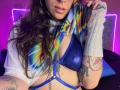 AmySpider with colorful tie-dye shirt, blue leather bra, and chain, showing tattoos, piercings, and lip ring, wearing gray wrist warmer, pink neon lights in background.