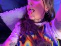 AmySpider with long brown hair, tongue out, metal chain in mouth, colorful tie-dye shirt, hoop earrings, tattoo on arm, purple and blue lighting, white fluffy pillow.