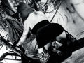 AmySpider's tattooed back and butt are exposed, wearing a black skirt and fishnet stockings, crouching with a tree branch in the background, in a gritty black-and-white photo.