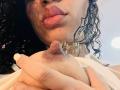 TiaraKitty's curly black hair, pink lips, and tan skin are in the foreground. She's holding her small, dark brown nipple with her right hand.