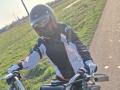 FrancaiseAnnaDom wearing a black helmet with a visor, red and black motorcycle jacket, white gloves, and black pants, riding a motorcycle on a grassy roadside.