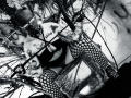 AmySpider with tattoos, wearing a black mask, fishnet stockings, and a glittery top, sits on the ground, holding a stick, with her big tits and ass visible.