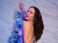 MollyMarmalade with long brown hair, topless, looking back, arms raised, showing small tattoos, blue-tinged skin, near a glittery Christmas tree, wearing a gold necklace.
