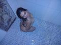 NatashaMonttero, naked, kneeling in a tiled shower, water dripping from her wet black hair, small black thong, smiling up, blue-tinted, with her ass and tits visible.