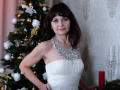 AngieGreen with dark brown hair, purple eyeshadow, white strapless dress, and a big jeweled necklace stands in front of a Christmas tree, smiling slightly. Boobs visible.