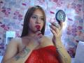 BelletinaXx with red lipstick, holding a black lipstick cap, wearing a red sparkly dress, has tattoos on her arm and hand, brown hair, and a silver bracelet.