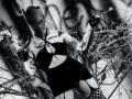 AmySpider with tattoos, wearing a black mask, strapless black top, and fishnet stockings, holding a cigarette. Her large boobs and ass are visible. She's surrounded by branches.