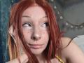 HannaCool with red hair, freckles, and piercings on nose and lower lip, looking sideways, holding her hair. Her yellow top is visible. Her eyes are slightly up.