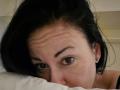 GennyRock's face is partially hidden in a white pillow, with dark black hair, brown eyes, and a small, gold hoop earring on her right ear. She has a worried expression.