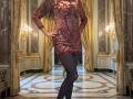 MaitresseElsa in a sheer red lace dress with sequins, black pantyhose, and long blonde hair, standing with one hand on her hip, showing her tits and ass.