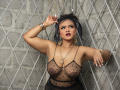 AliceJelanni, a curvy woman with tan skin, stands against a diamond-patterned wall. She wears a sheer black top revealing her large breasts, with gold jewelry and dark hair. Seductive, confident pose.