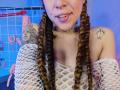 HellenK with braided hair, white fishnet top, nose ring, and tattoos, showing her tits, tongue out, and a choker, standing in front of BDSM gear.