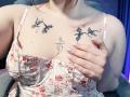 Rubbi with pale skin, floral dress, and tattoos on her chest. She's holding one large tit, showing black star and dragon tattoos, with her pink-manicured hand.