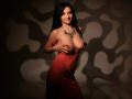 MadameAlexaX with black hair, red dress, and necklace, holding her big, bare boobs, smiling, in shadowy light. Her hand covers one tit, showing her nipple.