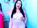 EmelySofia with long black hair, pink off-shoulder top showing small tits, and pink panties, sticking out her tongue, sitting in a blue-lit room, holding her top.