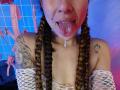 HellenK has braided hair, a nose ring, and her tongue out with saliva dripping. She's wearing white mesh sleeves, showing tattoos on her shoulders, and has small breasts.