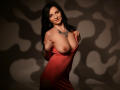 MadameAlexaX with large, bare tits, wearing a low-cut, red dress, holding it up, black wavy hair, necklace, standing, dark shadow pattern, confident, sexy, warm lighting.