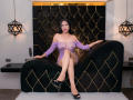 AgathaPember with black hair, purple sheer top exposing her tits, black high heels, sitting on a black and gold patterned chair, legs crossed, looking at camera.