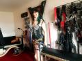 MaitresseElsa stands in a room with BDSM gear, wearing a black, studded, cleavage-revealing dress and a colorful belt, showing her tits and ass, with green-tipped hair.