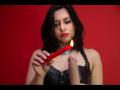 Maitresse Belle, com cabelo escuro, batom vermelho, blusa de couro preta, segurando uma vela vermelha, chama pequena, expressão séria, colar preto, fundo vermelho..