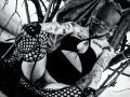 AmySpider with tattoos, wearing fishnet stockings, black bodysuit, and glasses, has dreads, black gloves, and boots, posing with her legs up, showing her pussy and ass. Black-and-white photo.