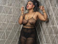 AliceJelanni, sexy, dark-skinned woman with black hair in ponytail, wearing sheer black polka dot dress, exposing breasts and nipples. Gold jewelry, standing against diamond-patterned wall, seductive pose, holding cigarette.