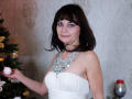 AngieGreen with medium tits, wearing a white strapless dress and a chunky silver necklace, holding a Christmas ornament, has dark brown hair and purple eyeshadow, smiling.