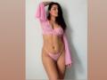 HeidyMillerr with dark hair, pink sheer top, and pink thong, showing her tits and pussy, has one arm raised, standing with a slight smile, and a confident pose.