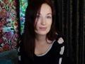MarleneDavies has pale skin, blue eyes, and dark red hair. She's wearing a black top with white straps, smiling slightly, and looking at the camera.
