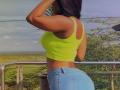 KatyBomb with tan skin, long dark hair, wearing a neon yellow crop top, showing her boobs, and tight blue jeans, standing on a balcony, hands in her hair.