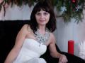 AngieGreen with medium tits, wearing a white strapless dress and silver necklace, sitting on a black fur couch, dark brown hair, purple eyeshadow, smiling, Christmas decorations in background.
