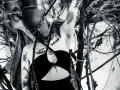 AmySpider with shaved head, tattoos, glasses, black strapless top, chain necklace, and arm raised, standing among tree branches. Black-and-white image. Boobs, tattoos, and arm tattoos visible.