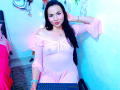 EmelySofia with long black hair, pink off-shoulder dress, visible nipples, sitting at a laptop, blue light, smiling, hand in hair, pink bed, pink wall, blue light.