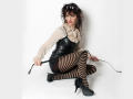 MaitresseElsa, with striped black-and-brown stockings, sits on the floor, wearing a black leather mini-dress, sheer white top, and black heels, holding a black strap.