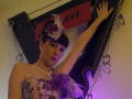 MaitresseElsa with dark hair, purple eyes, and lipstick, wearing a lace dress with flowers, raising arm on a black, studded, leather frame with chains and a red sign.