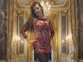 MaitresseElsa stands in an opulent room, wearing a sheer red dress with intricate patterns, black tights, and a confident smirk, hands on hips, brown hair flowing.