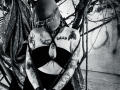 AmySpider with shaved head, glasses, black strapless top, large tits, arms covered in tattoos, wearing black choker, bound with ropes, black and white, gritty, sexy.