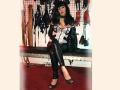 MaitresseElsa with black hair, wearing a white corset, black leather gloves, and pants, sitting on a bench, showing her tits, with black high heels and a red carpet.
