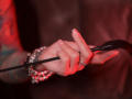 MissVonTease's tattooed hand grips a black riding crop, her manicured nails and diamond ring visible, with red lighting highlighting her wrist adorned with a silver chain bracelet.