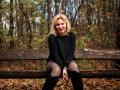 NatalyFoxy69 sitting on a wooden bench, wearing a black sweater, black tights, and black boots, with blonde hair and a smile, in a forest with fallen leaves.