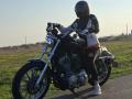 FrancaiseAnnaDom on a black Harley-Davidson, wearing a helmet, black jacket, and jeans, with a shiny helmet and boots, standing on a road, hands on the handlebars.